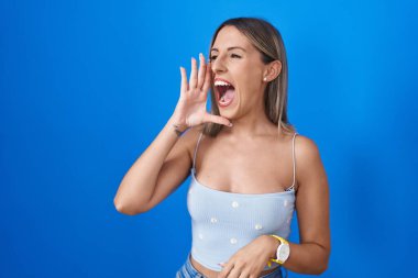 Young woman standing over blue background shouting and screaming loud to side with hand on mouth. communication concept. 