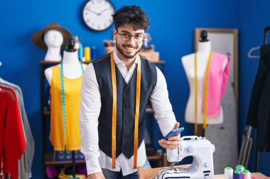 Young hispanic man tailor smiling confident using smartphone at sewing studio