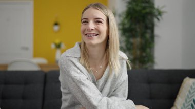 Young blonde woman smiling confident sitting on the sofa at home