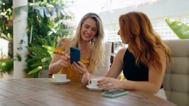 Akıllı telefon kullanan ve kahve içen iki kadın evdeki terasta oturuyor.