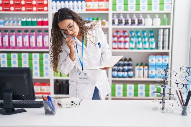 Genç, güzel İspanyol kadın eczacı telefonda eczanede defter okuyor.