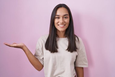Young hispanic woman standing over pink background smiling cheerful presenting and pointing with palm of hand looking at the camera. 