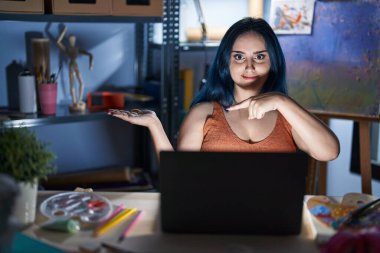 Mavi saçlı genç bir kız, geceleri dizüstü bilgisayarla resim stüdyosunda oturuyor kameraya hayret ediyor ve gülümsüyor.. 