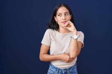 Mavi arka planda dikilen genç İspanyol kadın bir soru hakkında endişeli ve gergin bir şekilde düşünüyor. 