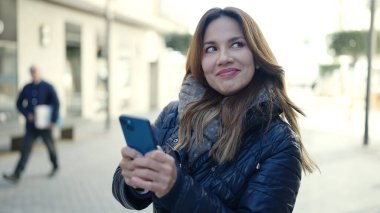 Genç, güzel, İspanyol bir kadın sokakta akıllı telefon kullanarak gülümsüyor.
