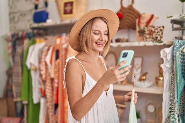 Young blonde woman customer wearing summer hat using smartphone at clothing store