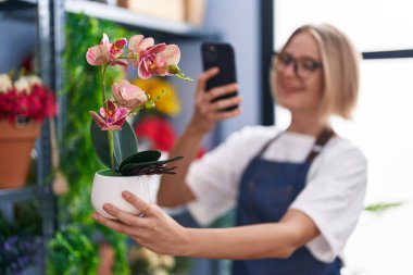 Genç sarışın kadın çiçekçi çiçekçiye çiçek dikmek için akıllı telefondan fotoğraf çekiyor.