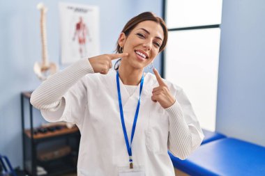 Acıyı dindirme kliniğinde çalışan esmer bir kadın neşeli bir şekilde gülümseyip parmaklarını ve ağzını işaret ediyor. diş sağlığı kavramı. 