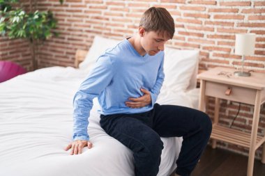 Young caucasian man suffering for stomach ache sitting on bed at bedroom