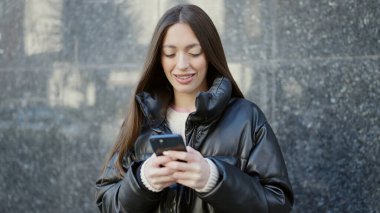 Genç, güzel, İspanyol bir kadın sokakta akıllı telefon kullanarak gülümsüyor.