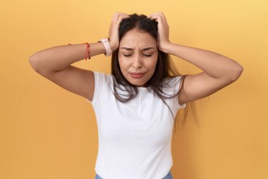 Sarı arka plan üzerine günlük beyaz tişört giyen genç Arap kadın acı ve migren yüzünden çaresiz ve stresli bir baş ağrısı çekiyor. Eller başının üstünde.. 