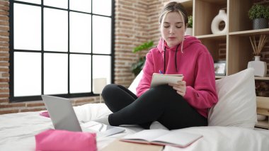 Young blonde woman student writing on notebook studying on bed at bedroom
