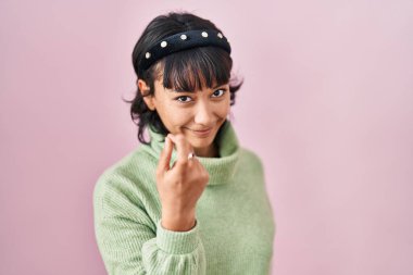 Young beautiful woman standing over pink background beckoning come here gesture with hand inviting welcoming happy and smiling 