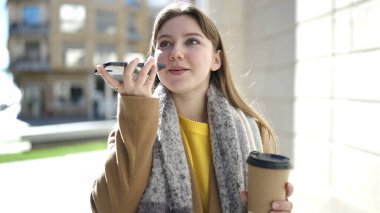 Young blonde woman talking on smartphone drinking coffee at street