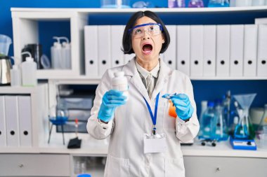 Young asian woman working at scientist laboratory angry and mad screaming frustrated and furious, shouting with anger looking up. 
