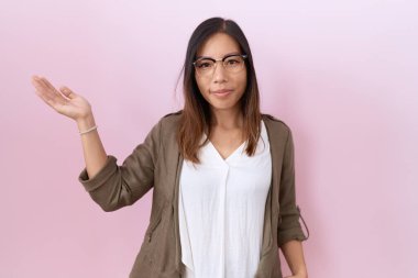 Orta yaşlı Çinli bir kadın pembe arka planda gözlük takıyor. Gülümsüyor, eli avuçlarında kameraya bakıyor.. 