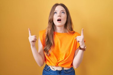 Caucasian woman standing over yellow background amazed and surprised looking up and pointing with fingers and raised arms. 