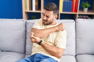 Young hispanic man sitting on sofa suffering for elbow pain at home