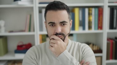 Young hispanic man student sitting on table with doubt expression at library university