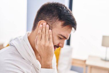 Young hispanic man suffering for ear pain sitting on bed at bedroom
