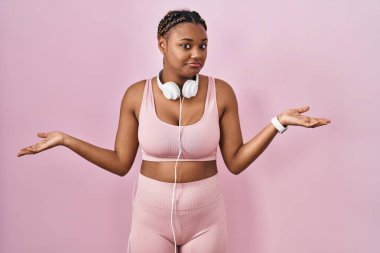 Spor kıyafetleri ve kulaklıkları takan Afrika kökenli Amerikalı kadın elleri ve kolları havada ve kafası karışmış bir ifade takınıyor. şüphe kavramı. 