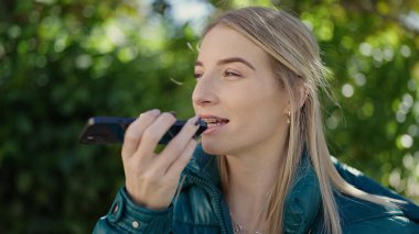 Young blonde woman sending voice message with smartphone at park