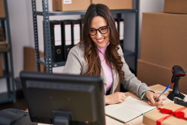 Young hispanic woman ecommerce business worker writing on notebook at office