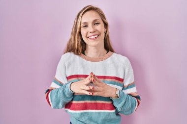 Young blonde woman standing over pink background hands together and fingers crossed smiling relaxed and cheerful. success and optimistic 