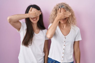 Anne ve kızı pembe arka planda yan yana durup gülümseyerek ve gülerek şaşkınlıktan gözleri kapatıyorlar. kör kavram. 