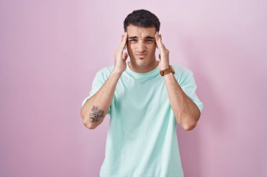 Pembe arka planda duran yakışıklı İspanyol adam, elleri başının üstünde, stres yüzünden başı ağrıyor. migren ağrısı. 