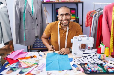 Young latin man tailor smiling confident cutting cloth at atelier