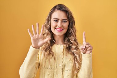 Sarı arka planda dikilen genç beyaz kadın altı numaralı parmaklarıyla gülümseyerek kendinden emin ve mutlu bir şekilde onu işaret ediyor.. 