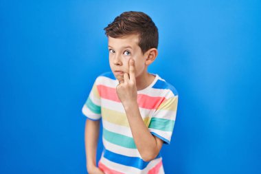 Mavi arka planda duran genç beyaz çocuk senin jestini ve şüpheli ifadeni izliyor. 