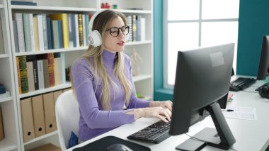 Young blonde woman student using computer and headphones studying at library university