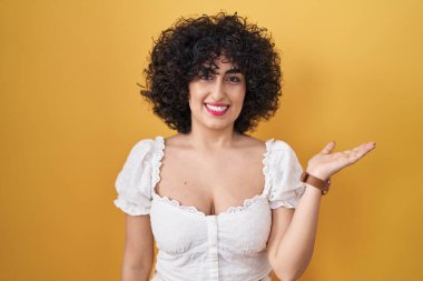 Sarı arka planda kıvırcık saçlı, genç esmer kadın gülümseyen, elinde avucuyla kameraya bakan.. 