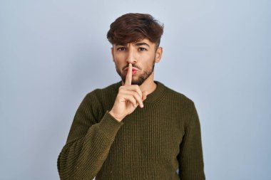 Arab man with beard standing over blue background asking to be quiet with finger on lips. silence and secret concept. 