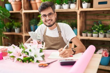 Young hispanic man florist counting dollars writing on notebook at flower shop