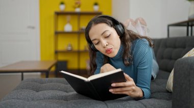 Genç ve güzel İspanyol kadın kitap okuyor. Kanepede uzanıyor ve kulaklık takıyor.