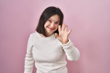 Down sendromlu bir kadın pembe arka planda durup mutlu, güler yüzlü, dostça bir jest yapıyor. 