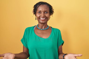 Sarı arka planda duran Afrikalı kadın. Kollarını açıp gülümsüyor. Dostça karşılama, olumlu ve kendinden emin selamlar. 