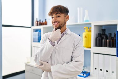 Laboratuvarda çalışan sakallı bir Arap adam kameraya gülümseyip kollarını kavuşturup çenesine bakıyor. Olumlu düşünmek. 