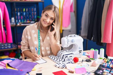 Young caucasian woman tailor talking on smartphone writing on notebook at sewing studio