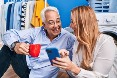 Orta yaşlı erkek ve kadın çift kahve içip akıllı telefon kullanarak çamaşır odasında çamaşır makinesini bekliyor.