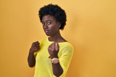 Afrikalı genç bir kadın sarı stüdyonun başında duruyor. Yumruk savunma hareketiyle dövüşmeye hazır. Kızgın ve üzgün bir yüz. 