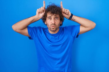 Mavi arka planda dikilen İspanyol genç adam, boynuz gibi parmağıyla komik jestler yapıyor. 