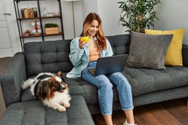 Genç beyaz kadın dizüstü bilgisayar kullanıyor. Kahve içiyor. Kanepede köpeğiyle oturuyor.