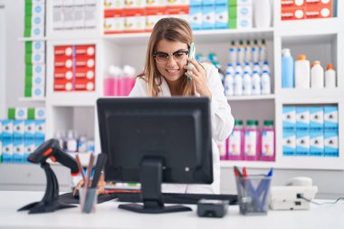 Young woman pharmacist talking on smartphone using computer at pharmacy