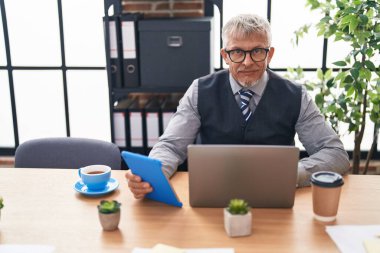 Ofiste dizüstü bilgisayar ve dokunmatik ped kullanan orta yaşlı, kır saçlı bir iş adamı.