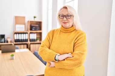 Orta yaşlı sarışın iş kadını. Ofiste kollarını kavuşturup el kol hareketi yapıyor.