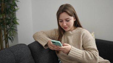 Evdeki kanepede oturan akıllı telefon kullanan genç sarışın kadın.